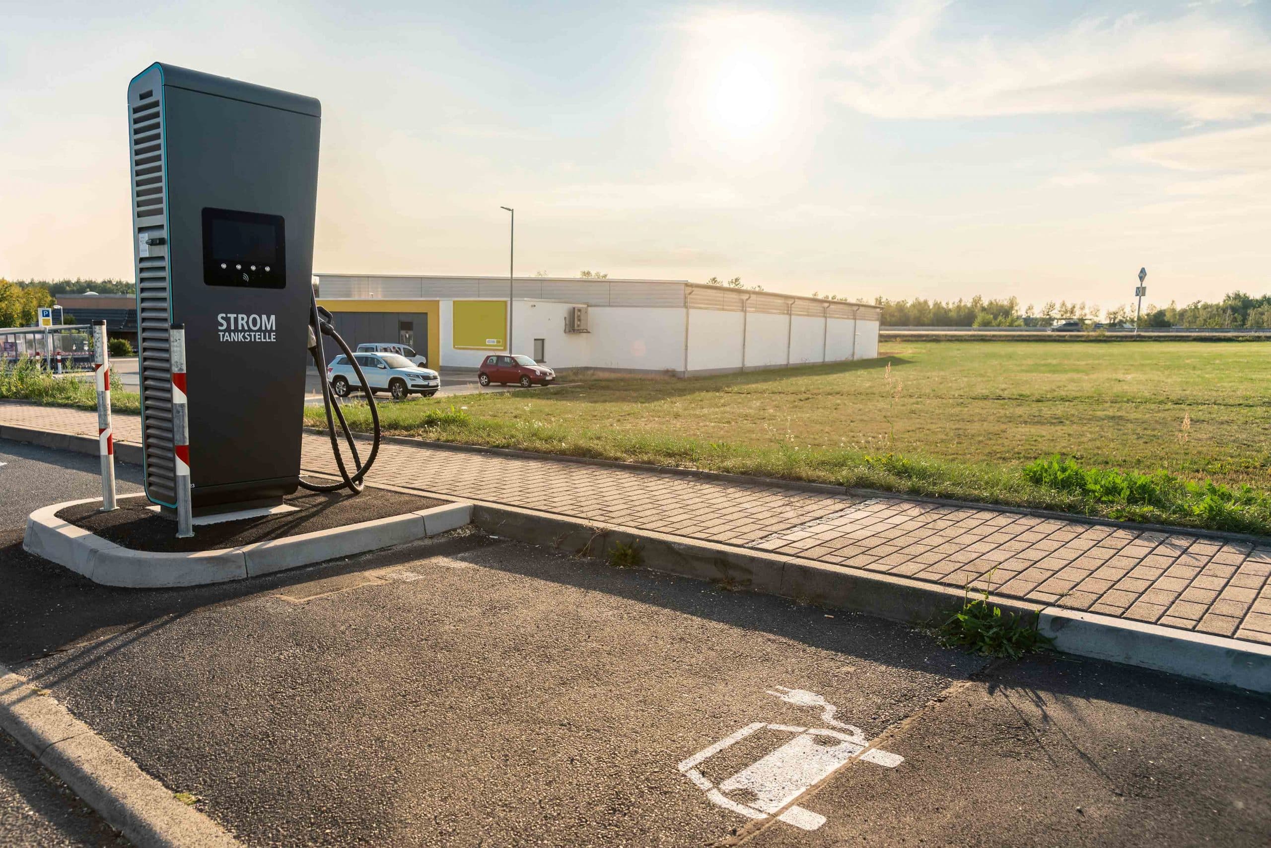 Ladestation für Elektrofahrzeuge auf Parkplatz neben Bürgersteig mit geparkten Autos und Gebäude.