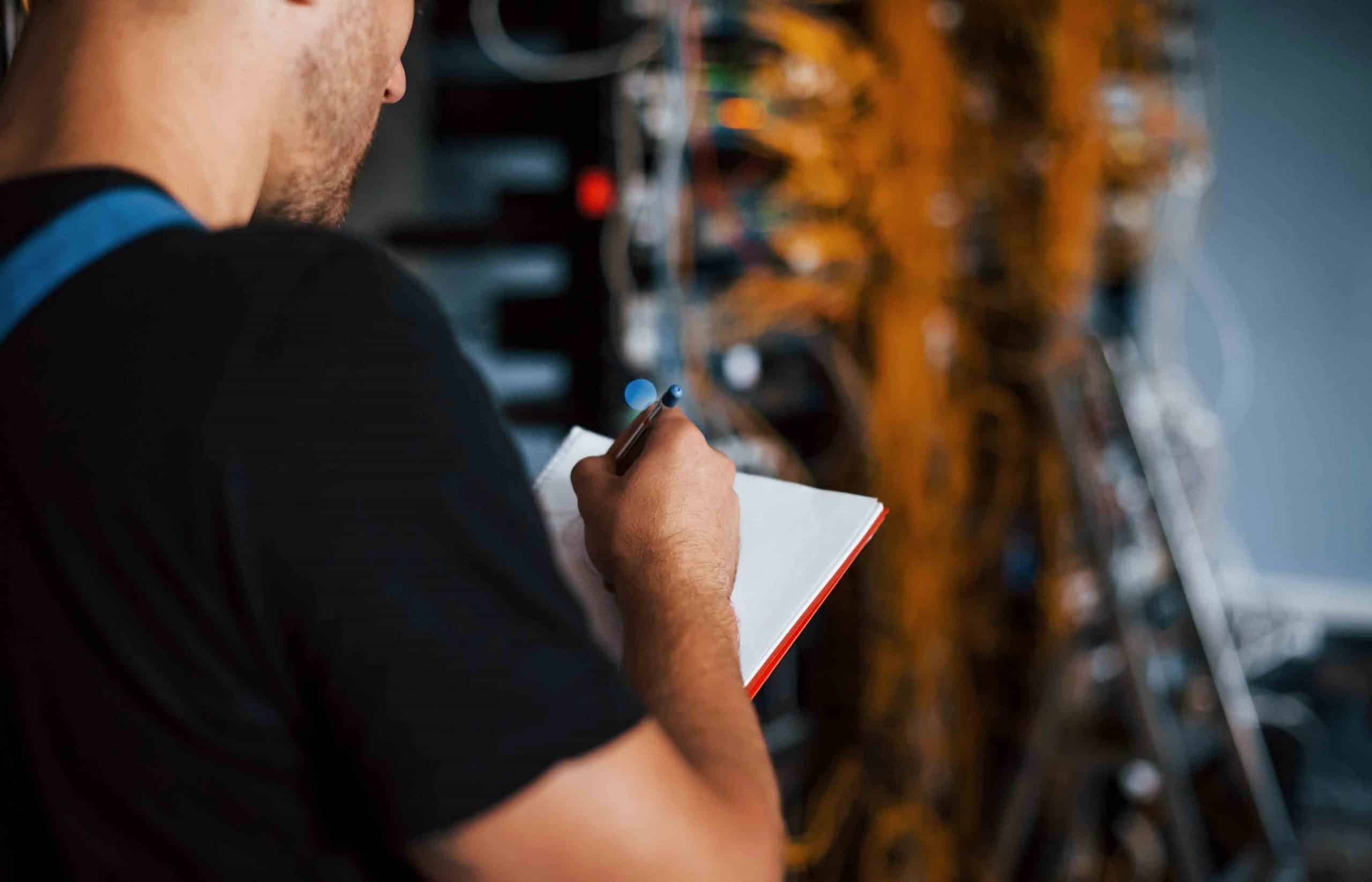 Mann notiert vor Netzwerkschrank mit zahlreichen orangefarbenen Kabeln.