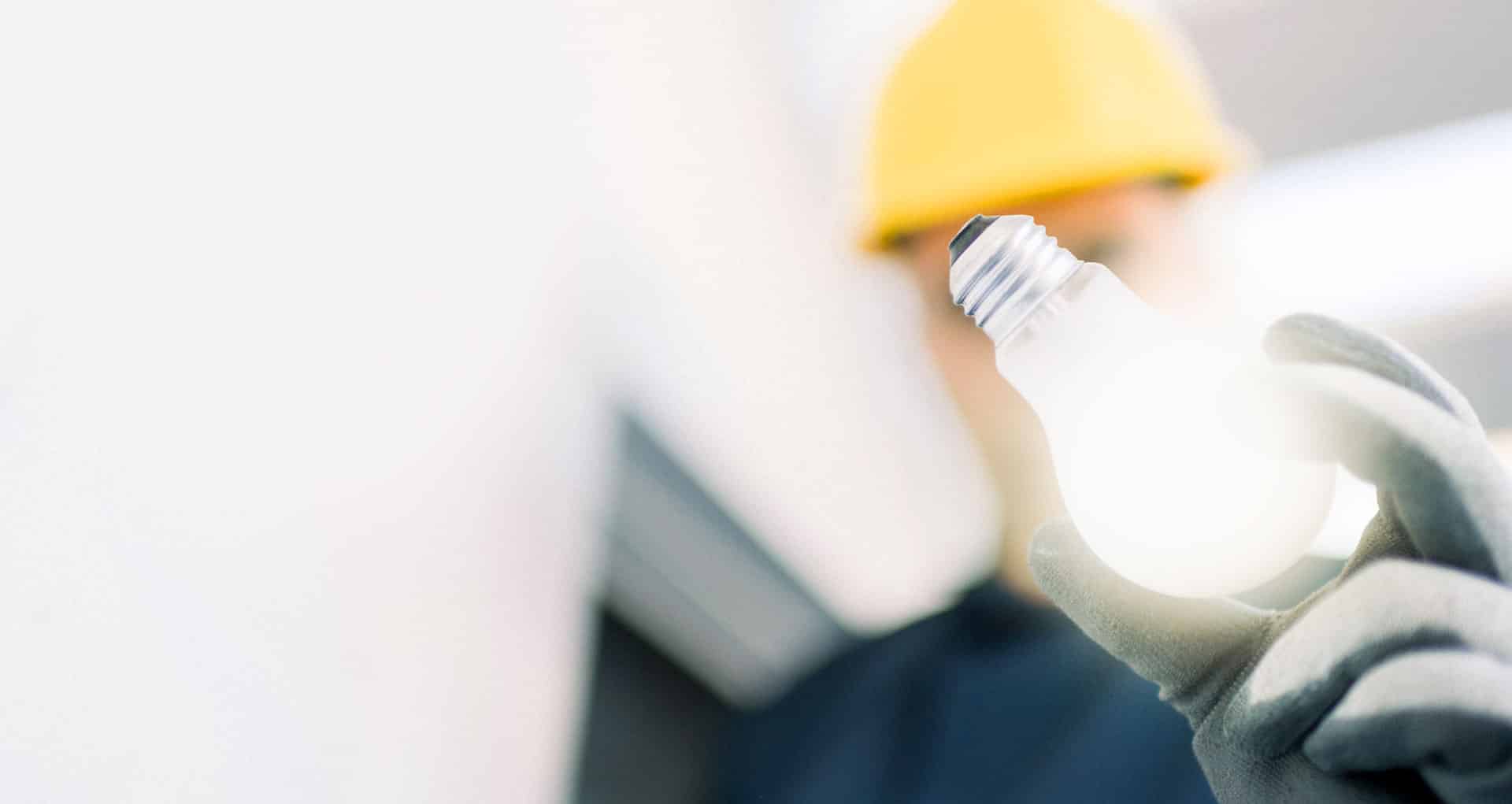 electrician_22 Person mit gelbem Helm und Handschuhen hält leuchtende LED-Glühbirne in Innenraum.