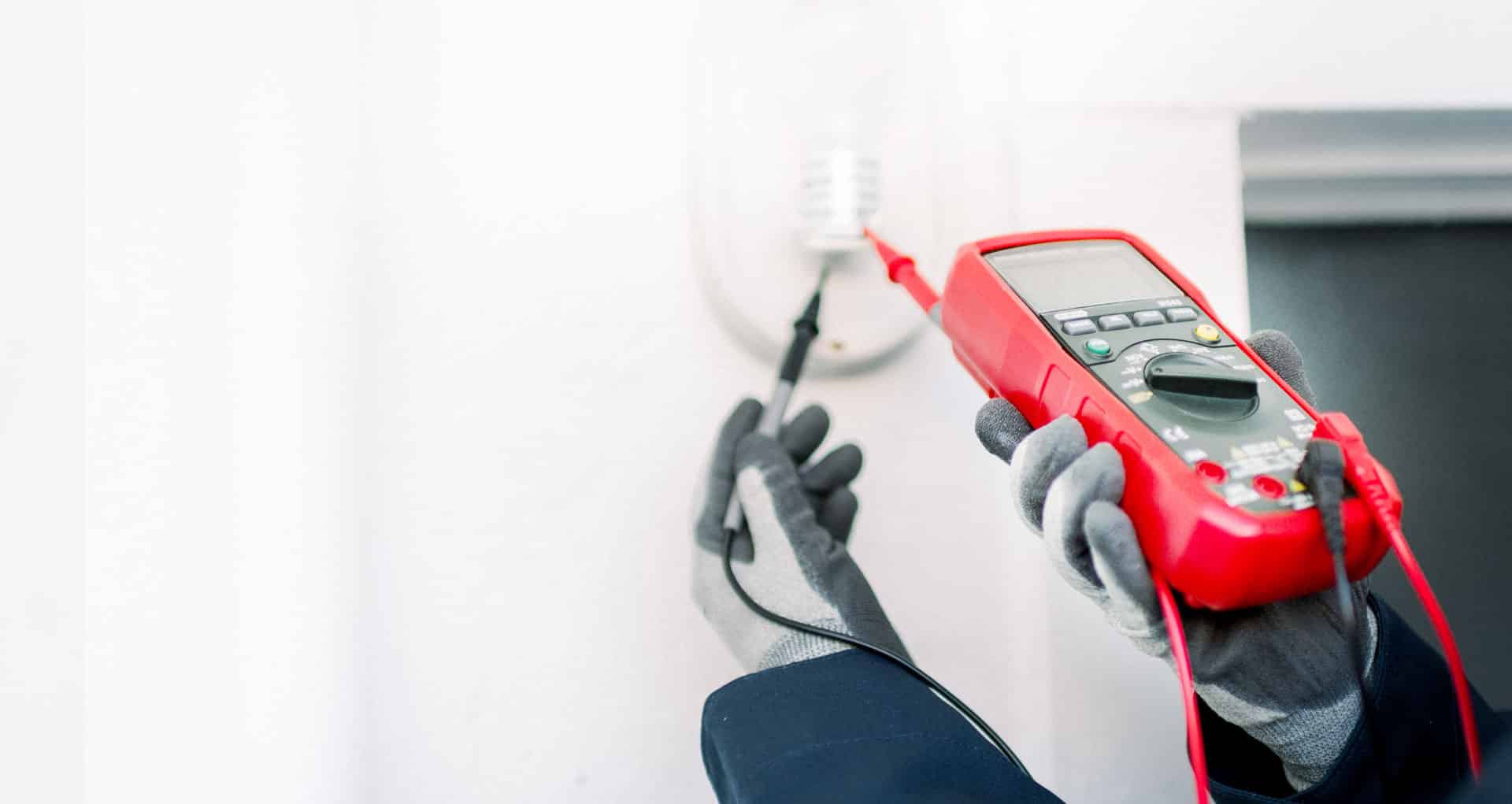 electrician_23 Person mit Handschuhen misst Strom in einer Wandsteckdose mit digitalem Multimeter.