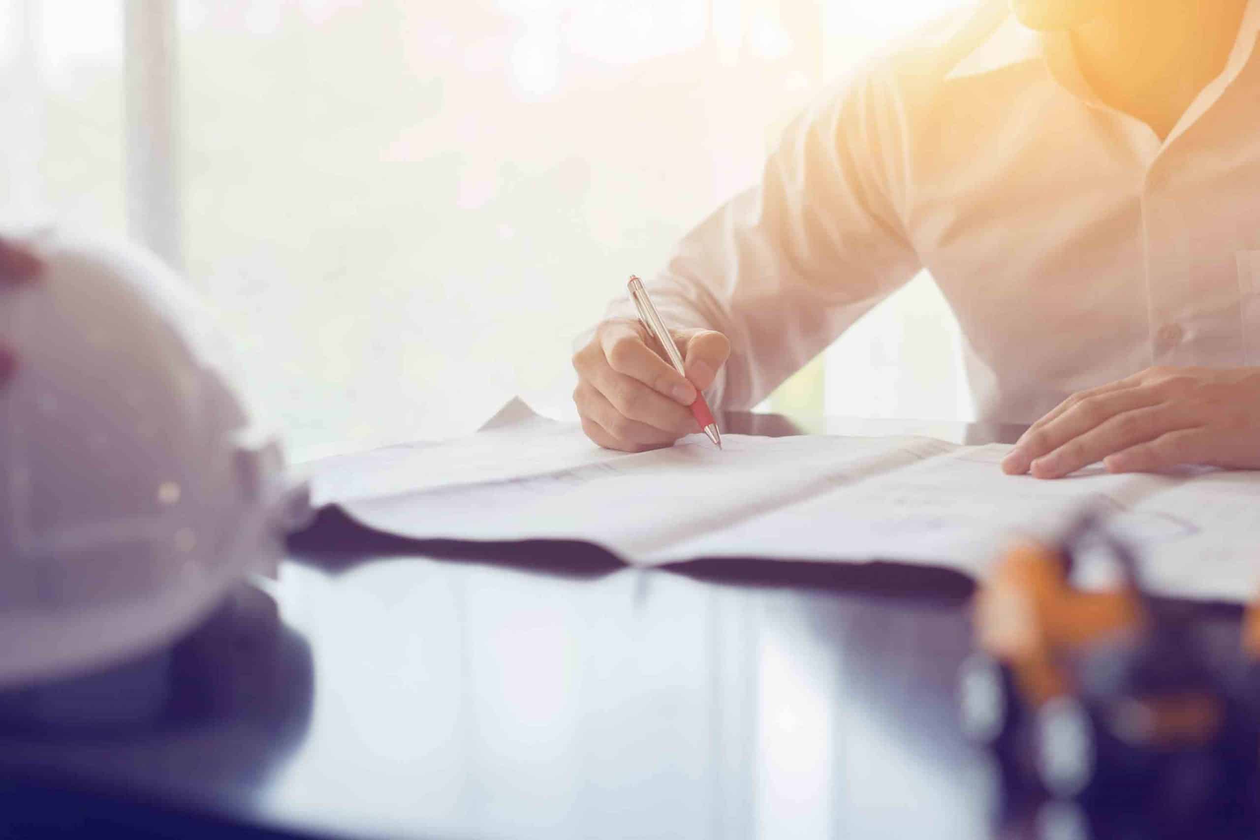 Person in weißem Hemd schreibt auf technische Pläne, weißer Schutzhelm liegt auf Tisch.