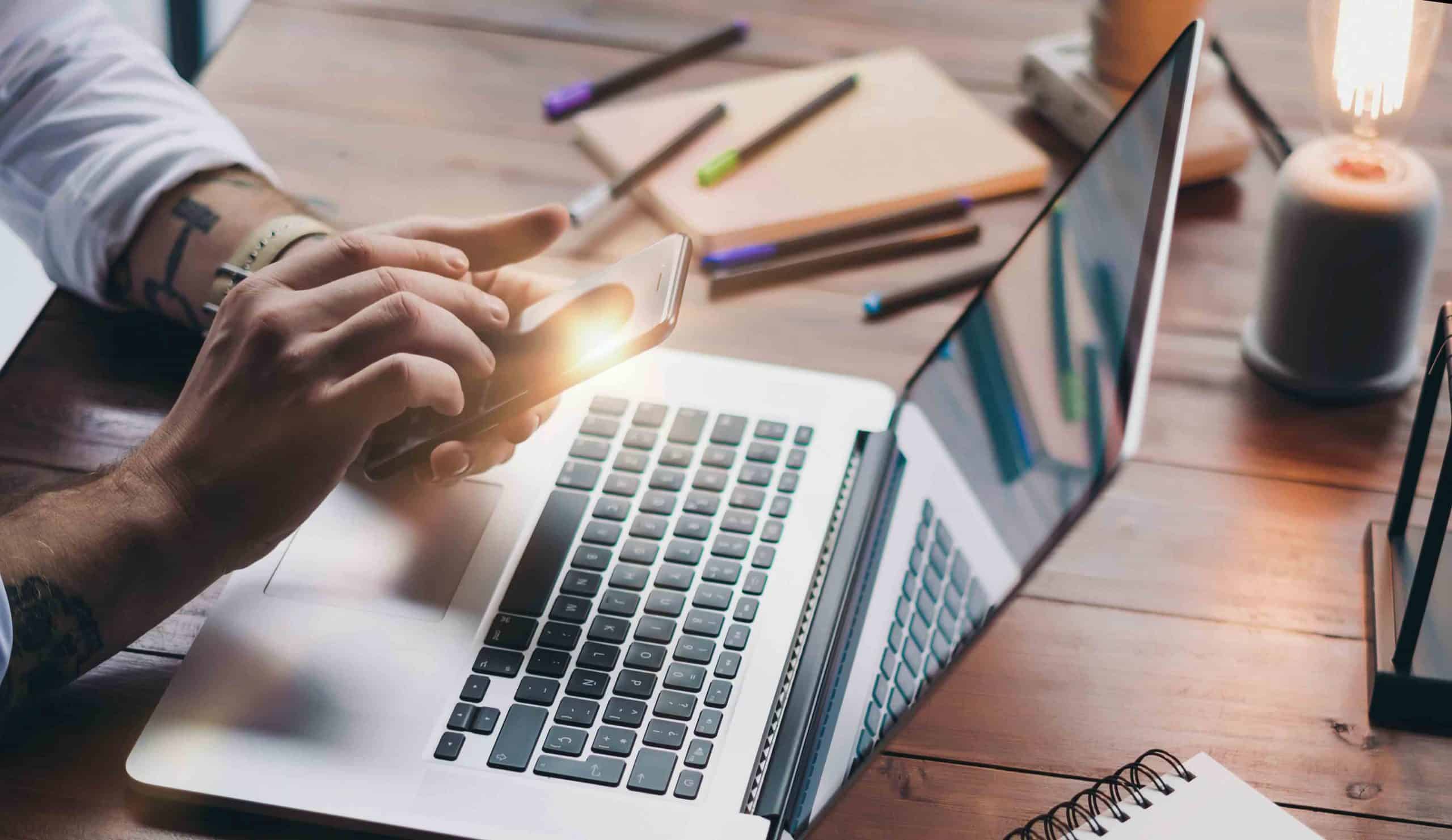 Person tippt auf Smartphone vor Laptop auf Holztisch mit Büroutensilien und Lampe.