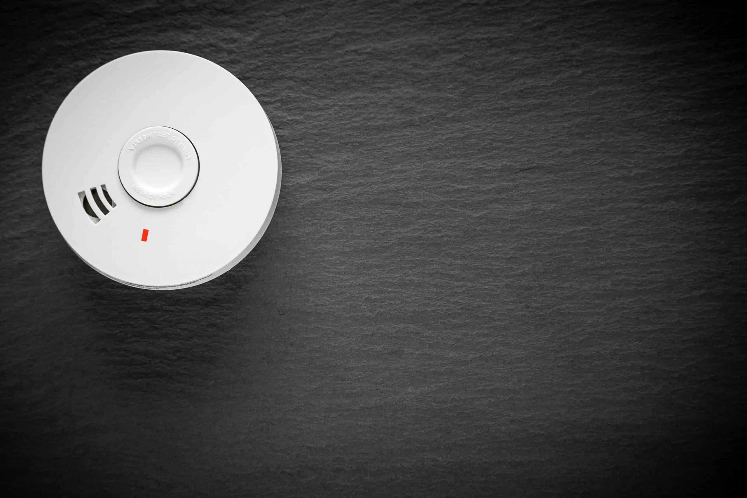 Weißer Rauchmelder an dunkler, strukturierter Wand montiert, frontal fotografiert.